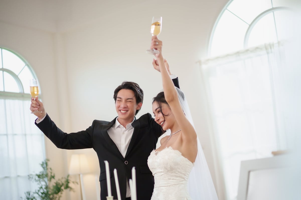 Happy Asian bride and groom celebrate wedding together with champagne toast inside studio, showing love and joy and raising glasses with happiness and cheers.