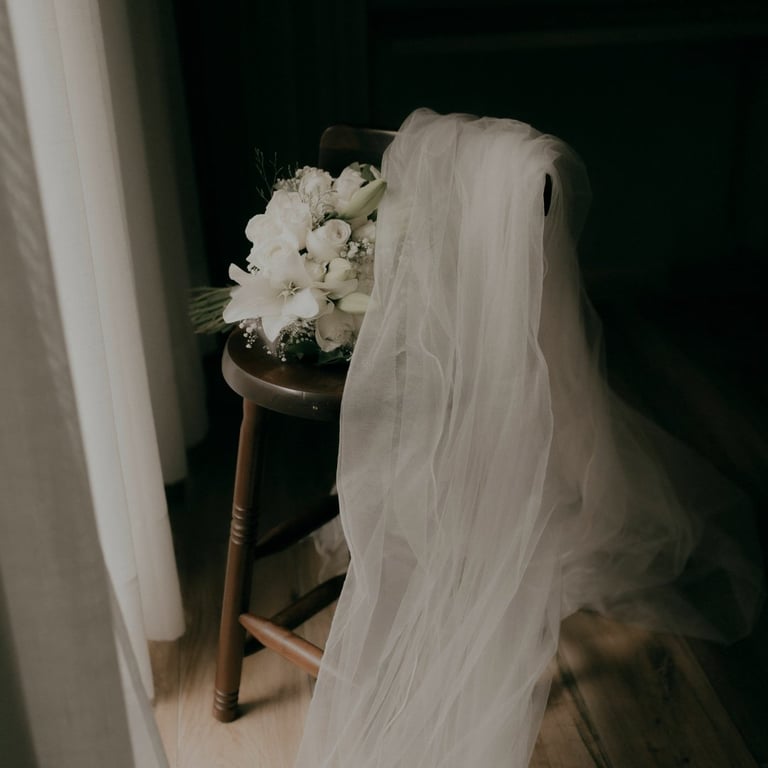 Bride from behind wearing long white veil next to wooden stool with white flower bouquet